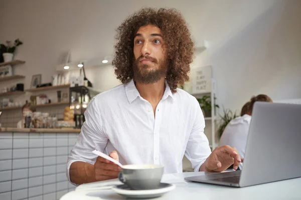 흰 셔츠를 입고 카페에서 테이블에 앉아 눈썹을 일으키고 이마를 수축, 놀랍게 도 멀리 보고, 원격으로 일하는 아름다운 젊은 턱수염 남자 — 스톡 사진