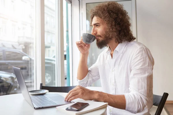 Young attractive man with long curly hair and beard sitting at table in cafe, working out of office with laptop, looking thoughtfully in window while having cup of tea — 스톡 사진