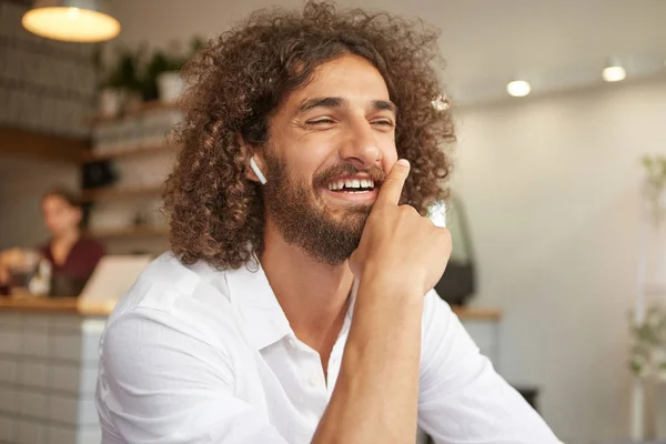 Indoor portrait of lovely curly bearded male in white shirt, smiling sincerely and leaning on the chin, having pleasant conversation in cafe, posing over indoor interior