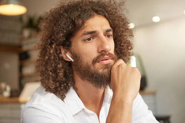 Puzzled attractive curly man with lush beard leaning on his chin and looking befogged forward, frowning and thinking about something important, posing over indoor interior