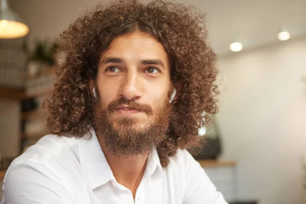Close-up portrait of attractive young male with beard, having long curly hair and brown kind eyes, having positive attitude, wearing white shirt
