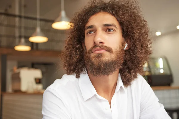 Photo en gros plan avec un beau mâle barbu en chemise blanche assis dans un café, regardant pensif de côté avec des écouteurs dans les oreilles, ayant pause déjeuner — Photo