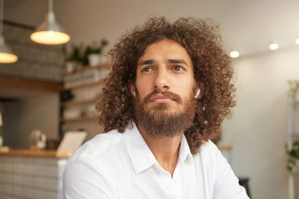 Closeup portrait of attractive curly guy with lush beard posing over cafe interior, wearing headphones and white shirt, looking aside pensively, waiting for his order — 스톡 사진