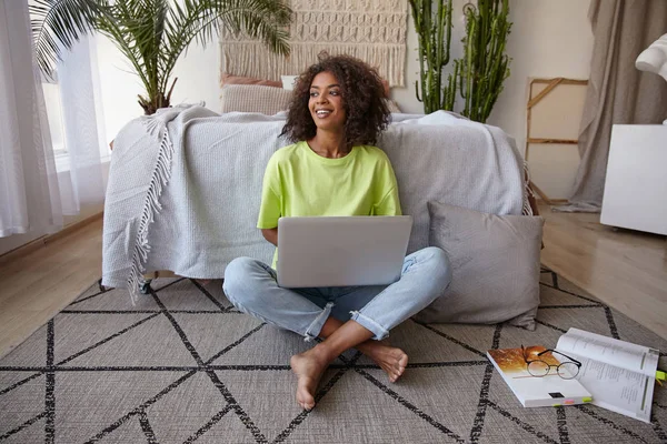 Cheerful young dark skinned woman sitting on floor in casual clothes, studying with texbooks and modern laptop, smiling joyfully and being in nice mood — 图库照片