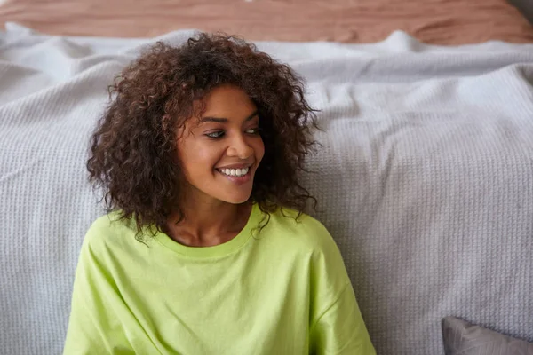 Primer plano interior de la joven mujer de piel oscura feliz con camiseta amarilla, posando sobre el acogedor interior de la casa, mirando a un lado con una sonrisa encantadora — Foto de Stock