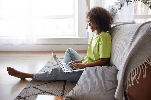 Atractiva mujer rizada joven sentada en el suelo con el ordenador portátil, manteniendo las manos en el teclado y mirando a la pantalla, con jeans y camiseta amarilla —  Fotos de Stock