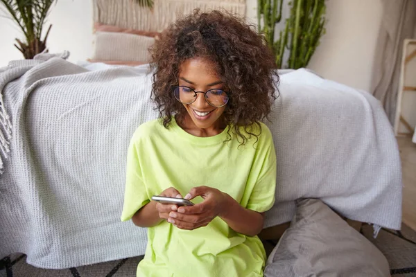 Happy young dark skinned curly female holding smartphone in hands, checking her social networks, looking at screen and smiling, posing over home interior