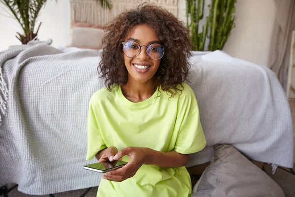 Retrato de encantadora joven feliz con piel oscura y rizos marrones, mirando la cámara alegremente con el teléfono inteligente en las manos, posando sobre el interior del hogar — Foto de Stock