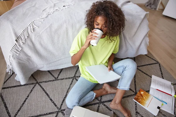 年轻的黑皮肤女性，棕色卷发，在家学习，坐在铺有几何图案的地毯上，喝咖啡，看笔记 — 图库照片