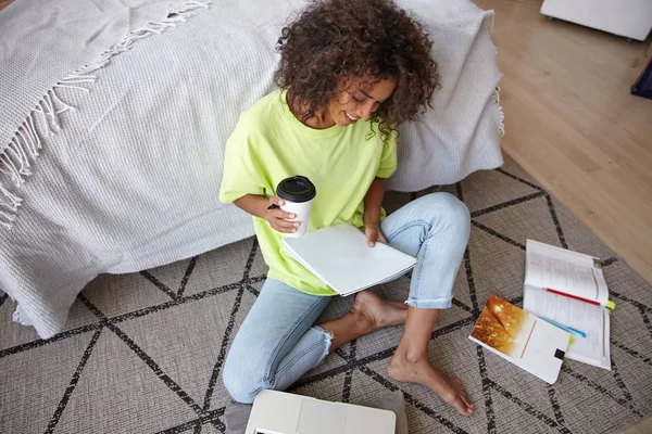 漂亮的年轻黑发卷发女子坐在地毯上，头戴几何图形，喝着咖啡，准备功课，穿着休闲装的室内照片 — 图库照片