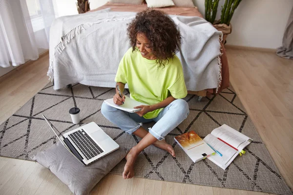Indoor shot of attractive dark skinned female with brown curly hair posing over home interior, preparing her homework with laptop, books and notes
