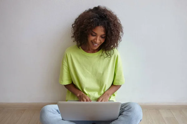 Retrato interior de una bonita joven con el pelo rizado marrón trabajando remotamente con el portátil, sentada en el suelo con las piernas cruzadas en ropa casual —  Fotos de Stock