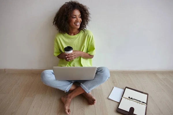 Happy beautiful dark skinned curly woman posing over home interior with cup of coffee in hand and laptop on legs, looking aside with broad smile