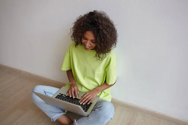 Foto interior de la joven mujer rizada feliz con la piel oscura charlando con amigos en el ordenador portátil, sentado con las piernas cruzadas, con pantalones vaqueros y camiseta amarilla —  Fotos de Stock