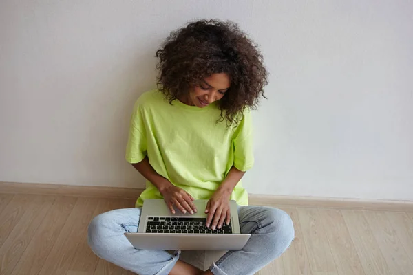 Joven bastante freelancer hembra con pelo rizado marrón sentado en el suelo con las piernas cruzadas, manteniendo el ordenador portátil en las piernas y trabajando en casa en ropa casual —  Fotos de Stock