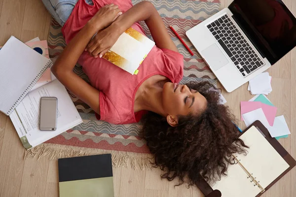 Hermosa joven rizada con la piel oscura tumbada en el suelo entre libros, cuadernos y portátil, posando sobre una alfombra de colores con los ojos cerrados y sonrisa agradable —  Fotos de Stock