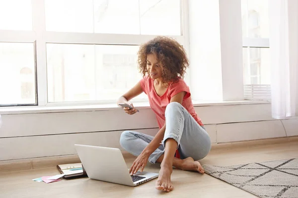 Foto interior de la encantadora mujer rizada de piel oscura sentada cerca de una gran ventana, sosteniendo el teléfono inteligente en la mano y revisando correos electrónicos en la computadora portátil, usando ropa casual. —  Fotos de Stock