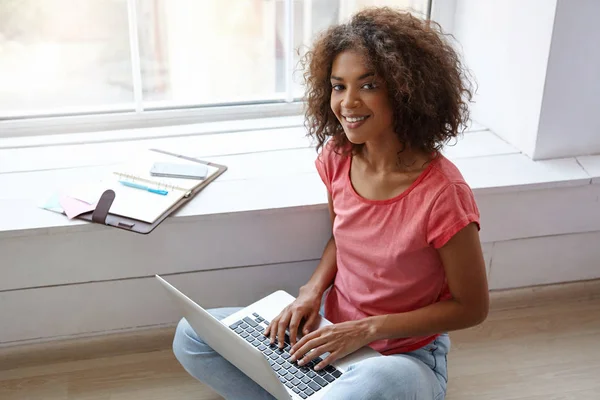 Indoor portrait of charming dark skinned woman with sincere smile posing over wide window on bright sunny day, working on windowsill with laptop and notebook