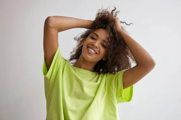 Primer plano interior de una joven positiva de piel oscura que mantiene su cabello con las manos, mirando a la cámara con una sonrisa encantadora, usando una camiseta amarilla — Foto de Stock