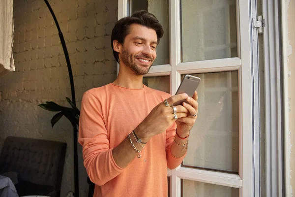 Photo d'intérieur d'un jeune homme brun positif en pull couleur pêche appuyé sur la fenêtre ouverte, tenant le téléphone portable dans les mains et regardant l'écran avec un large sourire — Photo
