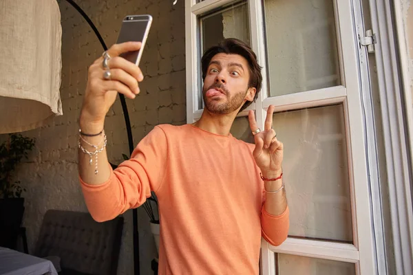 손에 전화기를 들고 창에 의지하고 우스꽝 스러운 얼굴을 하고 평화 표시를 하며 휴대폰으로 셀피를 찍고 있는 예쁘게 수염을 기른 남자의 이상 한 실내 사진 — 스톡 사진