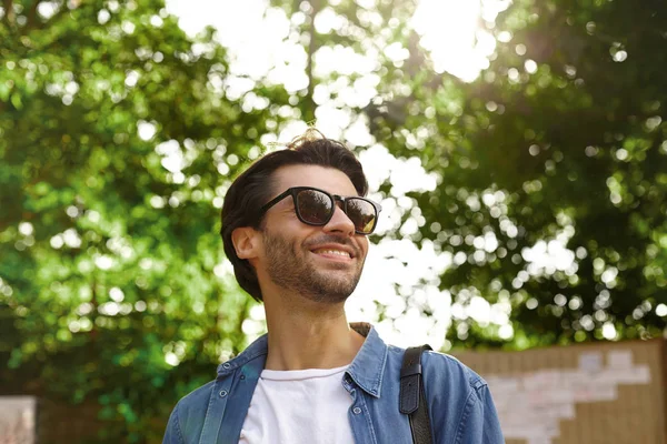 Portrait en plein air d'un joyeux jeune homme barbu aux cheveux foncés regardant de côté et souriant joyeusement, posant sur des arbres verts par une chaude journée lumineuse — Photo