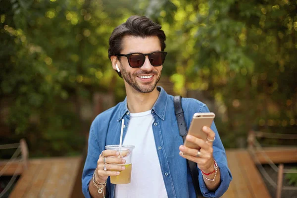 Portrait extérieur d'un beau mâle brun avec smartphone à la main, lisant de bonnes nouvelles et ayant une bonne humeur, buvant du thé glacé dans une tasse en plastique — Photo