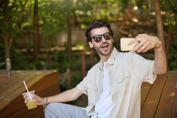 Portrait extérieur d'un beau jeune homme barbu faisant du selfie avec son smartphone sur un lieu public vert de la ville, regardant la caméra avec une large bouche ouverte, tenant une tasse de limonade à la main — Photo