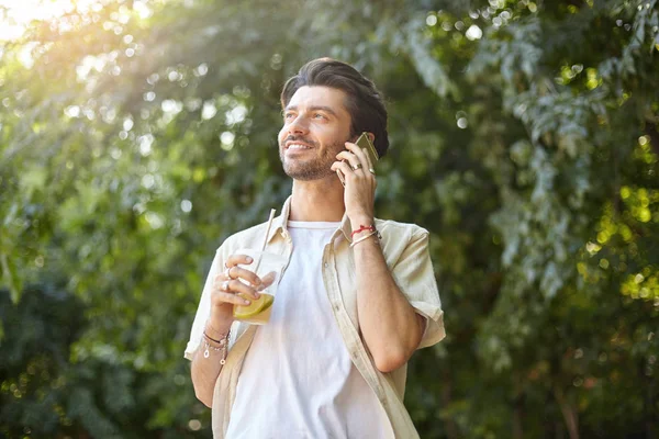 Joli jeune homme positif avec barbe posant sur le parc vert de la ville par une chaude journée ensoleillée, appelant avec son smartphone et tenant une tasse en plastique avec de la limonade — Photo