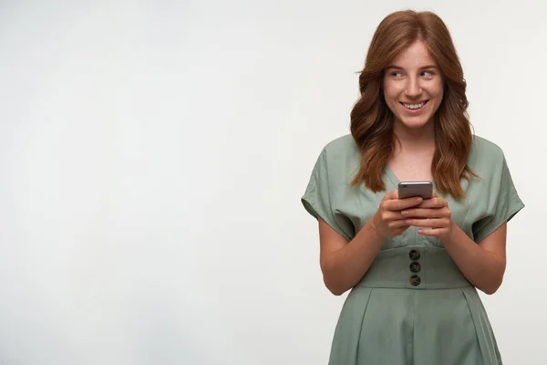 Gelukkig aantrekkelijk vrouw met rood haar poseren over witte achtergrond met mobiele telefoon in handen, opzij kijken en vrolijk glimlachen, het dragen van romantische jurk — Stockfoto