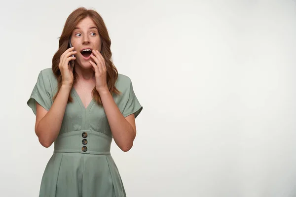 Sorprendido joven hermosa hembra con el pelo rojo posando sobre fondo blanco, abriendo ampliamente la boca y cubriéndolo con la mano, sosteniendo el teléfono inteligente por su oreja — Foto de Stock