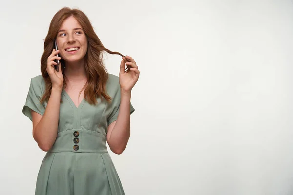 Porträt eines glücklichen rothaarigen Mädchens in romantischem Kleid, das zur Seite schaut und breit lächelt, telefoniert und die Haare am Finger verdreht, isoliert auf weißem Hintergrund — Stockfoto