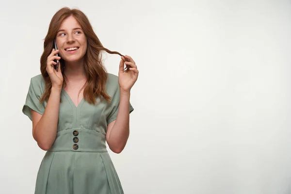 Ritratto di ragazza rossa felice in abito romantico guardando da parte e sorridendo ampiamente, parlando al telefono e torcendo i capelli al dito, isolato su sfondo bianco — Foto Stock