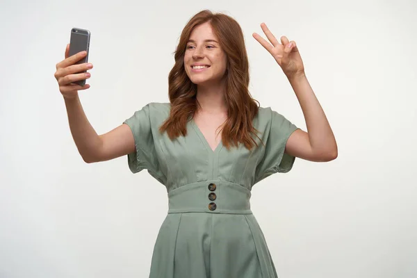 Retrato de una encantadora joven con una amplia sonrisa y un peinado romántico de pie sobre un fondo blanco, haciendo selfie en su teléfono móvil, mostrando un gesto de paz — Foto de Stock