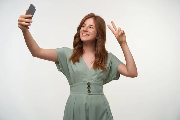 Estudio de toma de encantadora pelirroja joven hembra haciendo selfie con su smartphone, sonriendo a la cámara alegremente y levantando dos dedos con gesto de paz — Foto de Stock