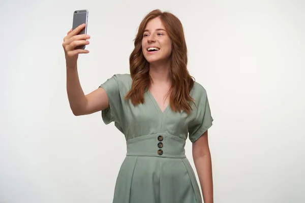 Indoor shot de alegre joven pelirroja posando sobre fondo blanco, haciendo una foto de sí misma con su teléfono, sonriendo a la cámara felizmente — Foto de Stock