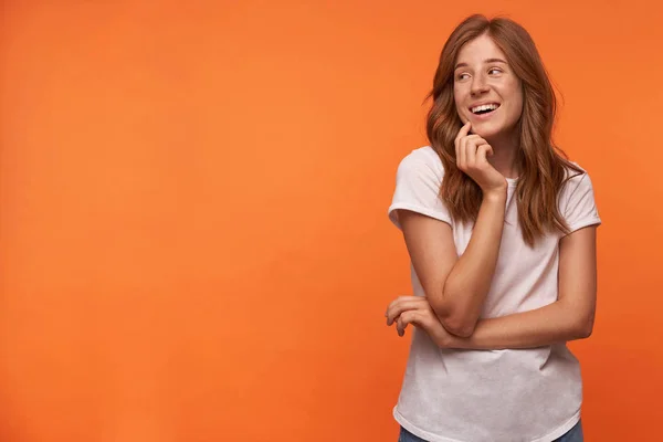 Estúdio tiro de linda jovem fêmea com cabelo vermelho segurando seu queixo com a mão, olhando para o lado e sorrindo alegremente, vestindo t-shirt casual — Fotografia de Stock
