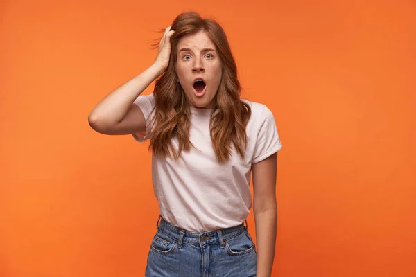 Foto de estudio de una joven sorprendida con ropa casual posando sobre fondo naranja con la boca abierta, sosteniendo su cabello y haciendo la cara de ojos abiertos — Foto de Stock