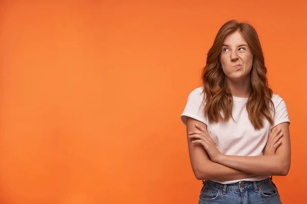 Foto interior de una joven atractiva posando sobre fondo naranja con la mano cruzada en el pecho, rizando los labios y mirando a un lado, usando ropa casual — Foto de Stock