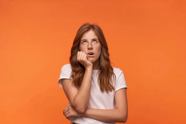 Studioaufnahme einer gelangweilten schönen Frau in weißem T-Shirt, die nach oben blickt, am Kinn angelehnt, mit trostlosem Gesicht, isoliert auf orangefarbenem Hintergrund — Stockfoto