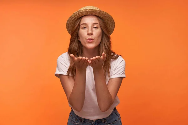 Estudio de toma de bastante rizado pelirroja mujer soplando aire beso a la cámara, levantando las palmas y doblando los labios en el aire beso, usando sombrero romántico y ropa casual — Foto de Stock