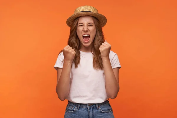 Retrato interior de una joven alegre con el pelo rojo levantando puños felizmente apretados, mirando a la cámara y gritando alegremente, posando sobre fondo naranja — Foto de Stock