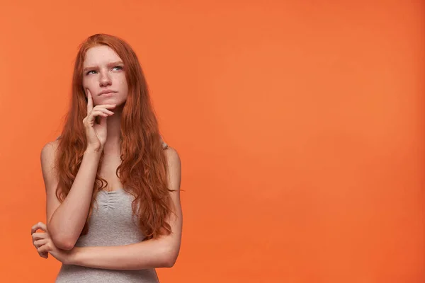 Bonito jovem mulher pensativa com cabelo aceno foxy cabelo loiro olhando para cima enquanto pensa, isolado sobre fundo laranja, vestindo camisa cinza casual, segurando a mão em seu rosto — Fotografia de Stock