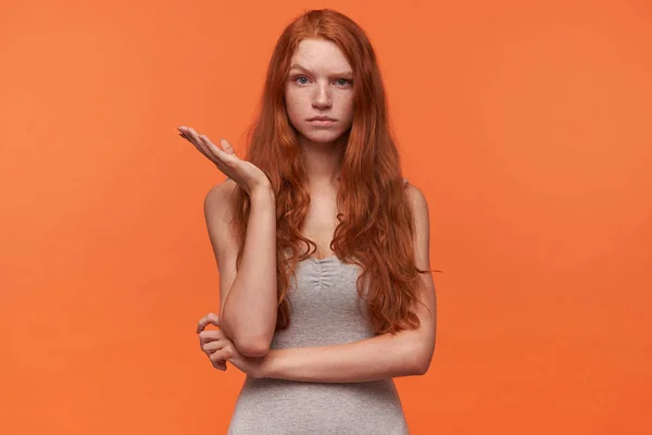 Attraktive junge rothaarige Frau mit welligen langen Haaren posiert vor orangefarbenem Hintergrund in lässiger Kleidung, blickt mit hochgezogener Augenbraue in die Kamera und hebt ratlos die Handfläche — Stockfoto