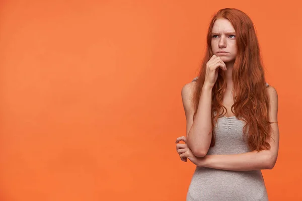 Horizontal portrait of young pretty woman with wavy foxy hair looking aside and frowning, having doubts in mind posing over orange background in casual grey shirt