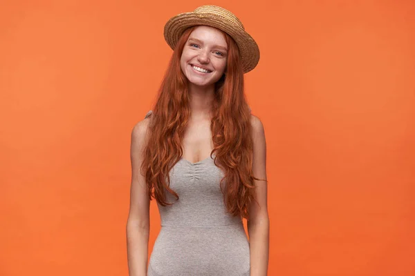 Horizontales Porträt einer angenehm aussehenden, glücklichen jungen Frau mit welligem Fuchshaar auf orangefarbenem Hintergrund, lässig gekleidet und mit Strohhut, Konzept positiver Emotionen — Stockfoto