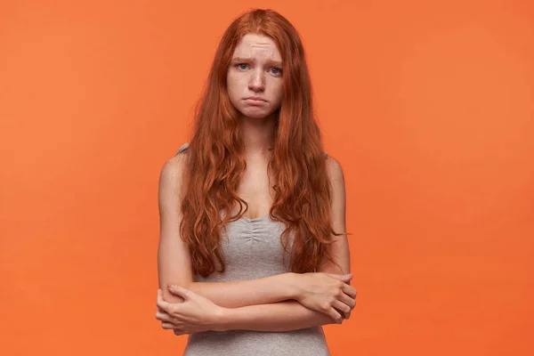 Beleidigte hübsche junge Frau mit welligem Fuchshaar posiert vor orangefarbenem Hintergrund, blickt mit traurigem Gesicht in die Kamera, trägt lässige Kleidung, hält die Hände gefaltet — Stockfoto