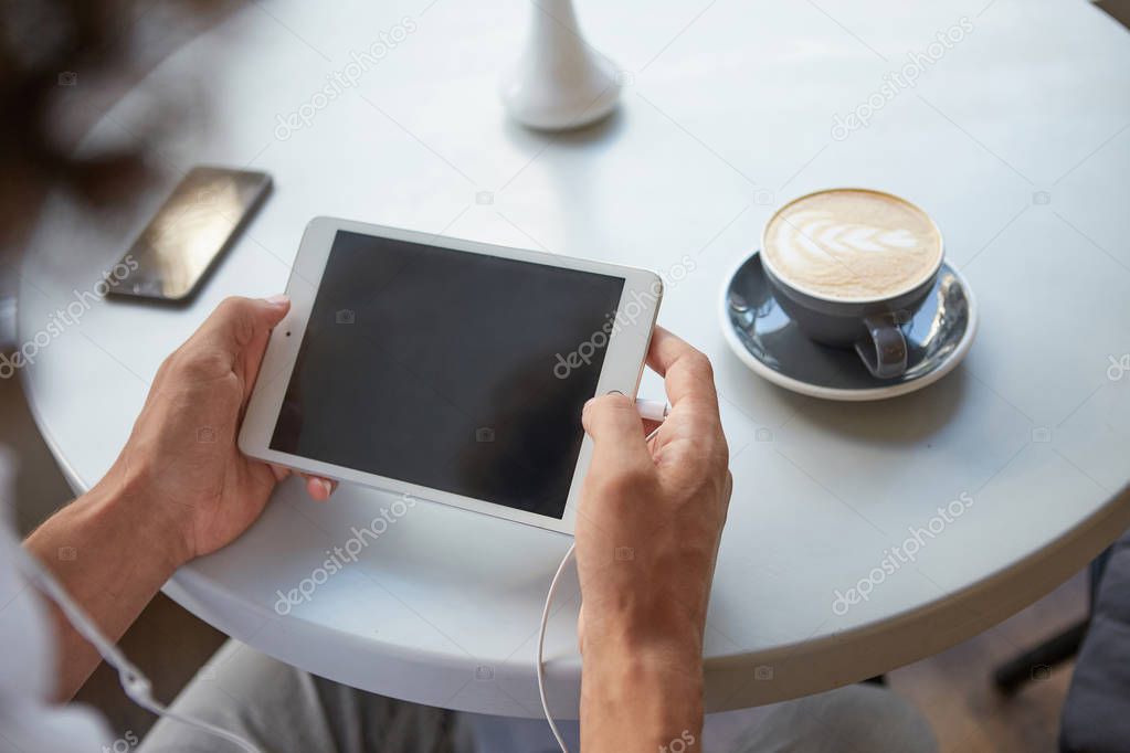 Indoor close-up of man's hands holding tablet with plugged earphones, having lunch break with cup of coffee, keeping smartphone on table