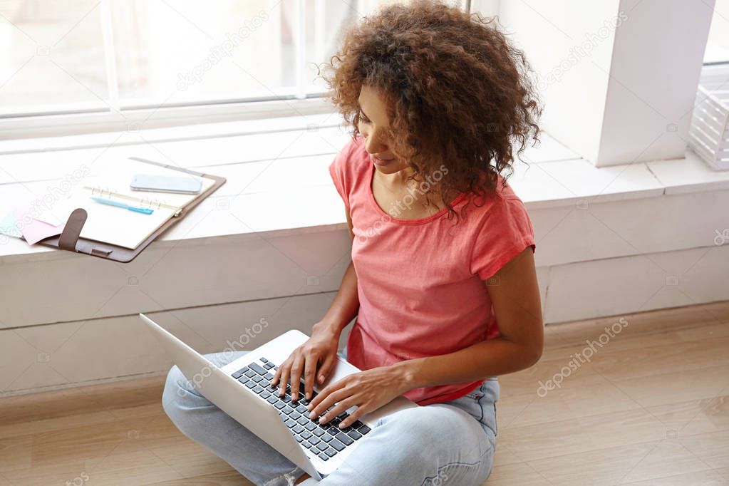 Portrait of young pretty curly woman with dark skin sitting on floor with laptop, keeping hands on keyboard, posing over wide window, wearing jeans and pink t-shirt