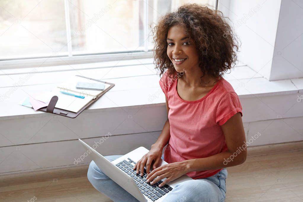 Indoor portrait of charming dark skinned woman with sincere smile posing over wide window on bright sunny day, working on windowsill with laptop and notebook